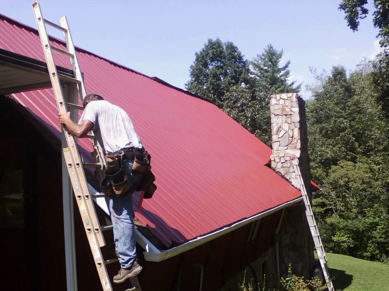 Roofer on a ladder beside a red metal roof and stone chimney — Soffit & Fascia Repair by Roofing Brothers in Demorest